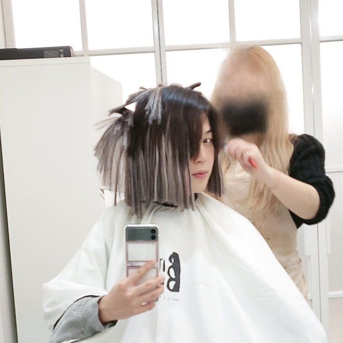 Jimin of BTS taking a picture with his phone while a hair stylist trims gray extensions that have been added to his black hair. One of his eyes is hidden behind long hair, and some hair is in clips on top of his head and is sticking out at odd angles. The untrimmed extensions hang down almost to his shoulders, making it look like he has a two-tone bob haircut, black on top and gray on the bottom.