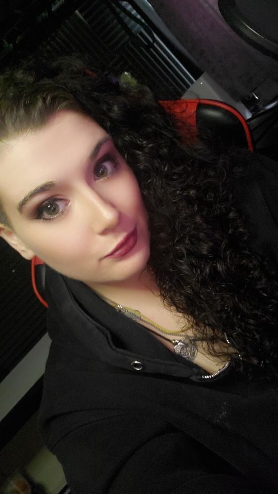 Photo of vala in her studio getting ready to stream.  She is wearing her long curly black hair down and she has on red violet lipstick and a smokey eye with black liner.  She is sitting in her red and black studio chair with her black hoodie on.