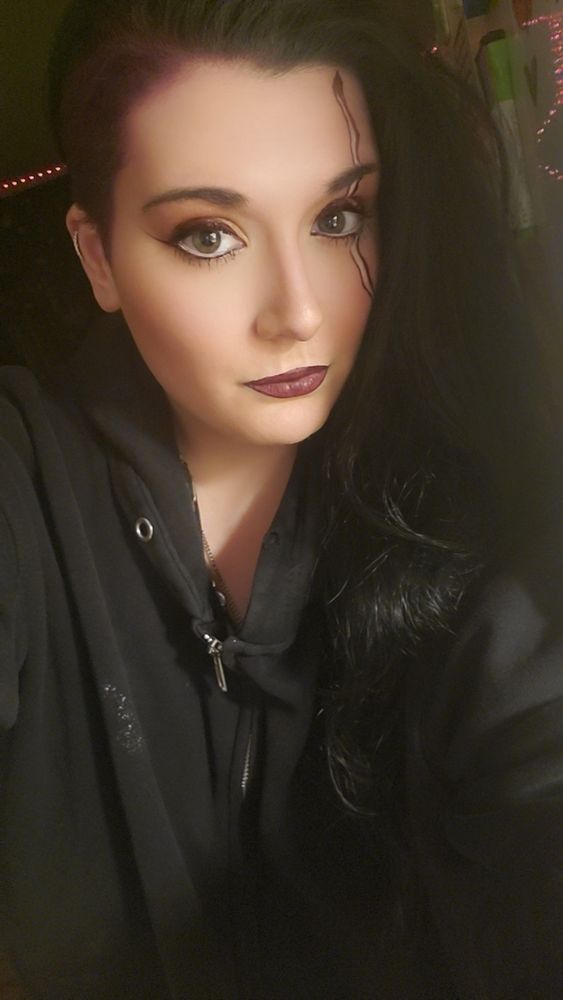 Photo of vala in what appears to be a different part of her house.  She has black and purple sideshaved hair, worn down and straight, and a black zipup hoodie on.  Her makeup is red, white, and purple, and appears to feature a long white and purple line extending downward across her face through her right eye.