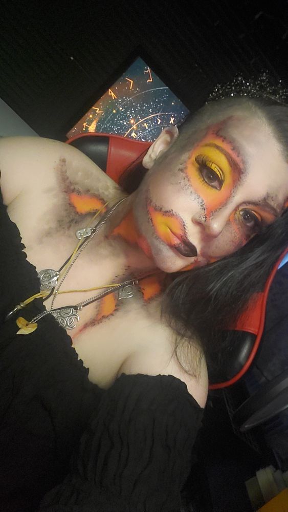 Photo of Vala wearing orange and black body art that resembles flames and falling ash.  She is wearing a black off the shoulder shirt and her black hair is long and straightened.  She has a crown on and she is in her studio.