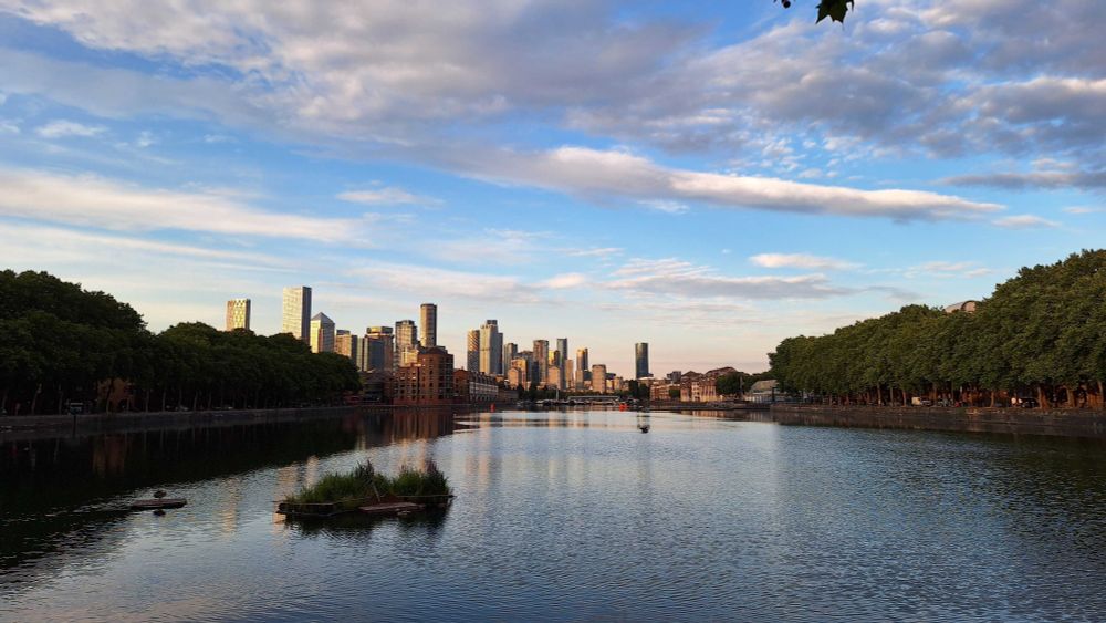A peaceful cityscape with a quay in the middle, tree-lined embankments on both sides, and modern skyscrapers in the distance, reflecting the golden light of sunset under a blue sky with scattered clouds.