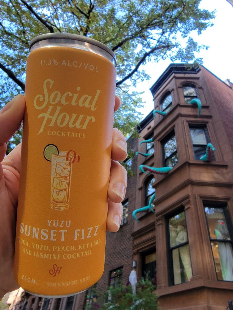 Aimée is holding a saffron colored can of Social Hour Cocktails Yuzu Sunset Fizz in front of building on the Upper West Side that was infiltrated by turquoise colored tentacles (at least nine are spotted outside of the windows in this Halloween display. )