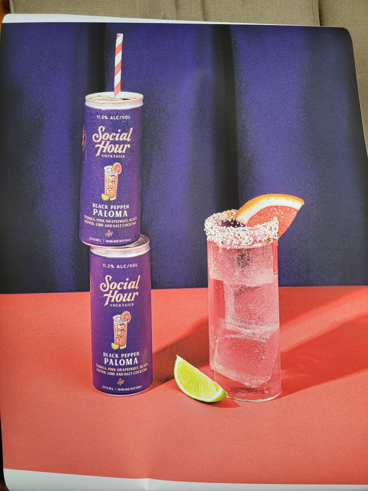 There is a purple can of Social Hour Cocktails Black Pepper Paloma with a red and white striped paper straw sticking out the top teetering on the shoulders of another can of Black Pepper Paloma like they ran off and joined the circus together.  To the right is a Black Pepper Paloma in a glass with a salted rim and a grapefruit garnish. A slice of lime lies between them without purpose.