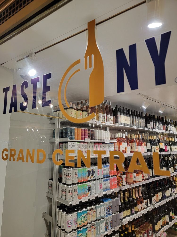 Image taken through the window of Taste NY in Grand Central of the display of several items including Social Hour Cocktails (Pacific Spritz, Yuzu Sunset Fizz, Whiskey Mule, and Navy Strength Gin and Tonic. )