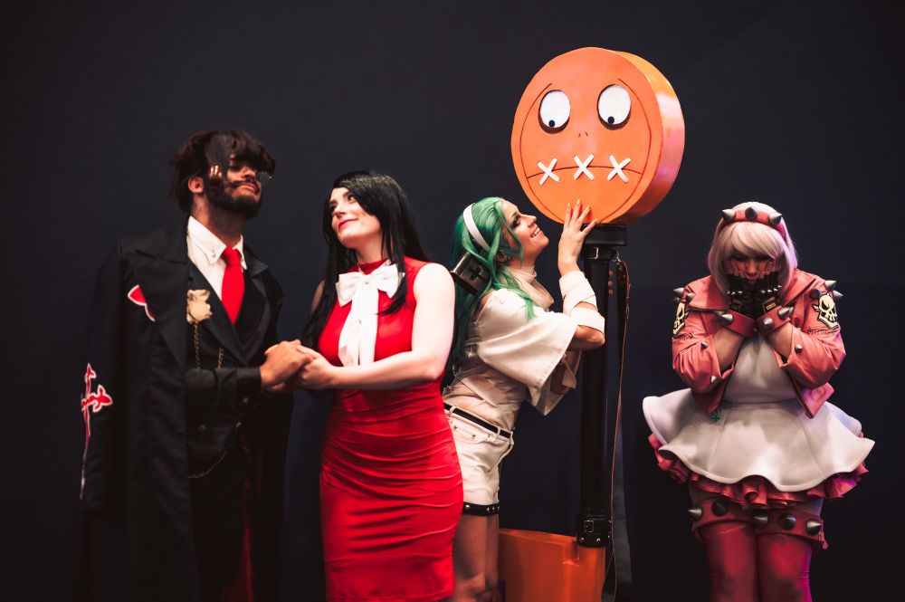 Maguma & princessbilbo lovingly holding hands while cosplaying as Slayer & Sharon, Steffvonschweetz cosplaying as A.B.A. with Paracelsus, staring up at him. & Piggynukka cosplaying as Elphelt, lamenting that she's not married like the others. Photo taken at EVO 2024