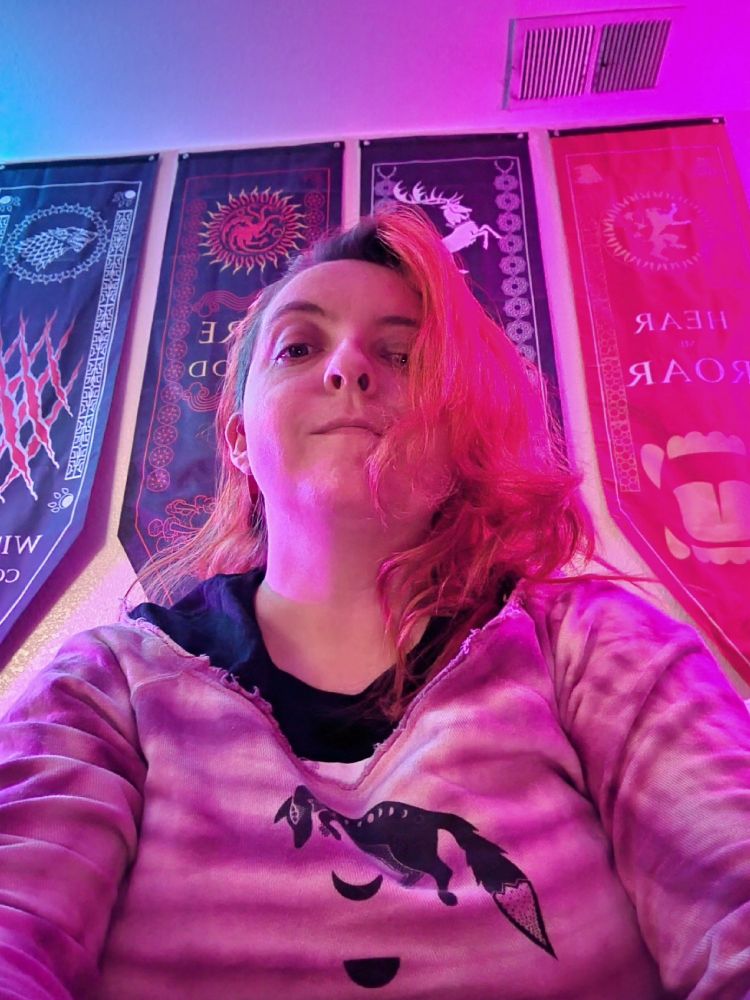 Myself, lit in teal and magenta light, red hair down, pink tie-dye sweater with a celestial fox and moons, over a black t-shirt, looking down at the camera, neutral expression. Several hanging pennants behind me.