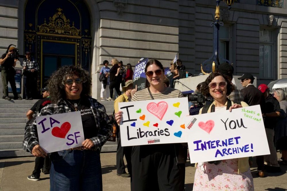 Three smiling people stand in front of San Francisco City Hall holding handmade signs celebrating the Internet Archive. Their signs read “IA ♥ SF,” “I ♥ Libraries,” and “♥ You Internet Archive.” Other participants and photographers are visible on the steps behind them.