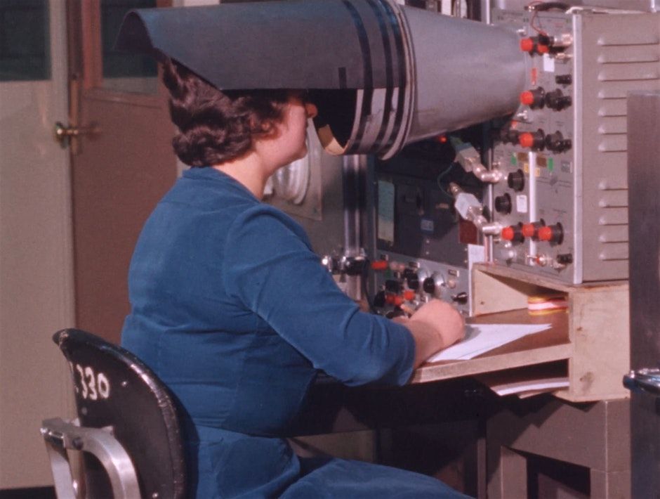 Single frame from a film from the Prelinger Archives. Depicts a woman taking notes while looking into the hooded scope of a piece of electronic equipment.