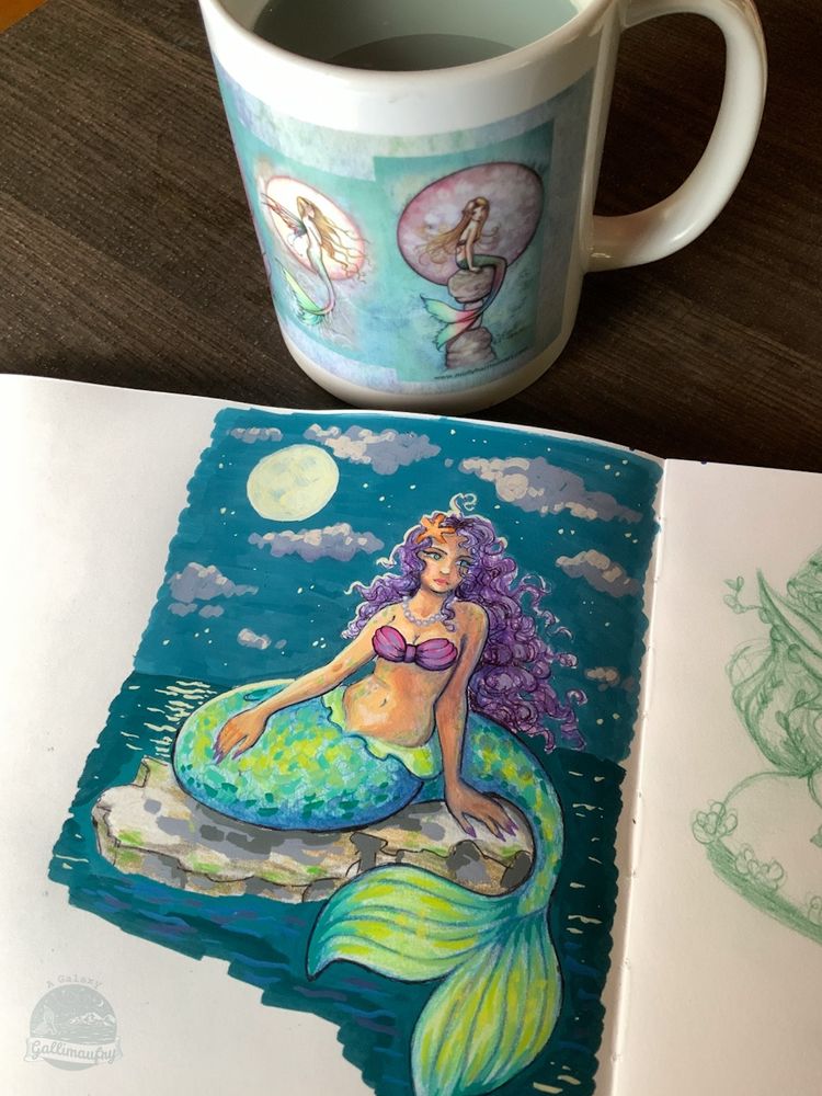 A photo of a cup of bluish-greenish colored tea and a mermaid drawing in a sketchbook. The mermaid is sitting on a rock in the middle of the ocean with the moon behind her in the night sky. The mug also has mermaid designs on it (the mug is not my design/artwork).
