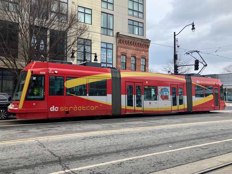 Photo looking across a city street to a streetcar vehicle. The vehicle is double-articulated, in predominantly red livery, and says "DC Streetcar". It is modern-looking and has a pantograph collecting current from the trolley wire.