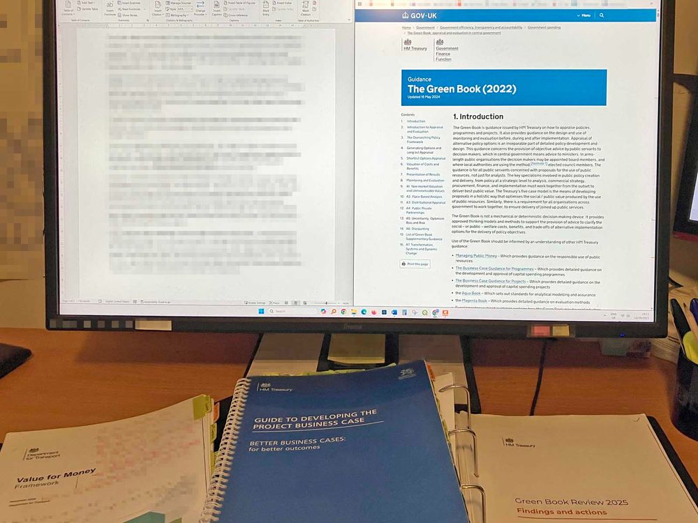 Photo of part of a desk, with a computer screen at the back. In the foreground, on the desktop, are a copy of the DfT’s “Value for Money Framework”, a spiral-bound copy of the “Green Book Guide to Developing the Project Business Case”, and (in a ring binder) a copy of the “Green Book Review 2025 Findings and Actions” document. The screen has two windows on it. On the right, there is the Green Book web page. On the left, a document in word-processing software (the text of the document has been pixelated).