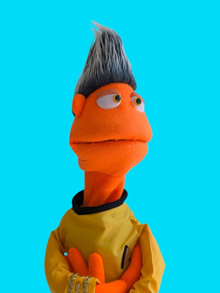 An orange puppet with a sky high peak of silver hair wearing a classic Star Trek inspired uniform.