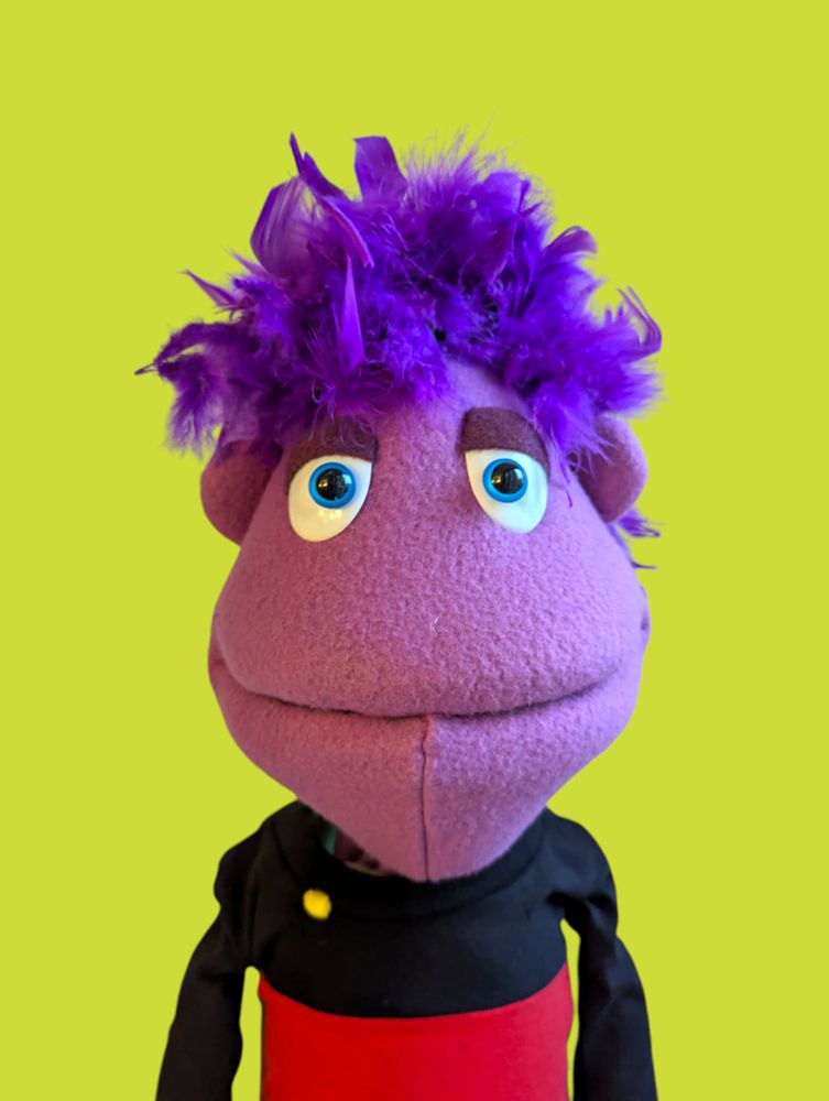 A purple puppet with feathery purple hair wearing a red and black uniform with a single pip.