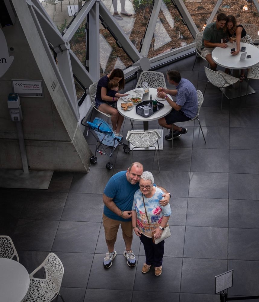 A man and an older woman stand closely together in the café area of the Dali Museum, smiling warmly as they look up toward the camera. They appear surrounded by modern geometric glass walls, tables, and visitors. The moment feels tender, capturing a special visit during an emotional time.