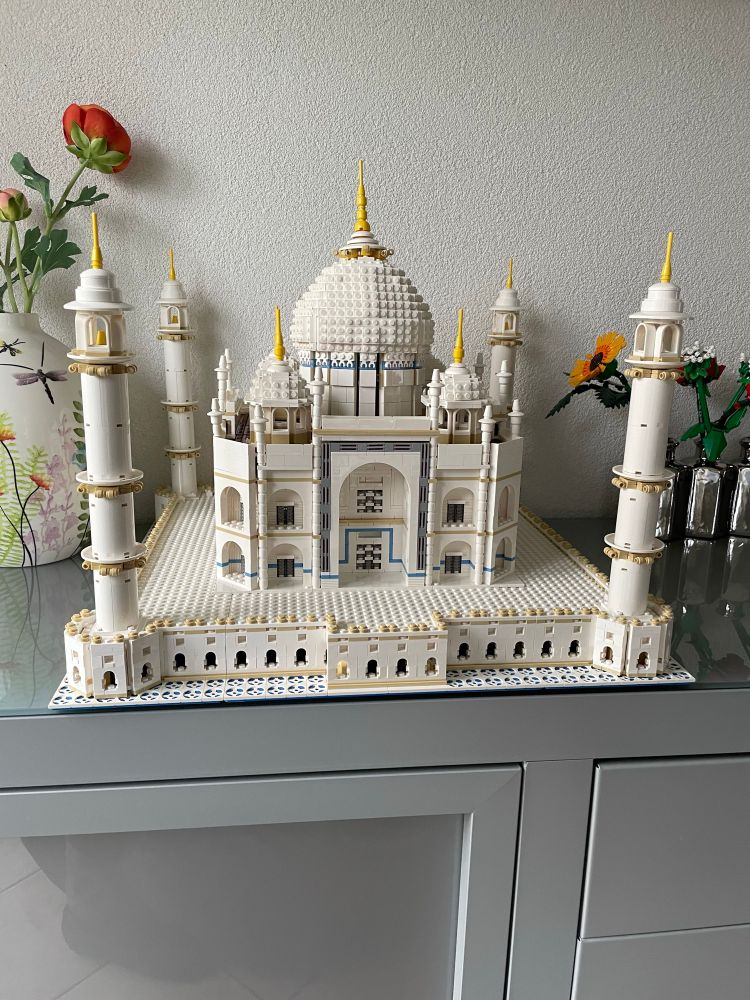 Een groot LEGO-model van de Taj Mahal staat op een kast. Het bouwwerk is voornamelijk wit, met gouden accenten op de torentjes en de koepel. Het model toont veel details zoals de bogen, ramen en de vier bekende minaretten op de hoeken. Op de achtergrond zijn een vaas met een rode bloem en wat andere bloemen van LEGO zichtbaar tegen een witte muur. De kast waarop het model staat is lichtgrijs.