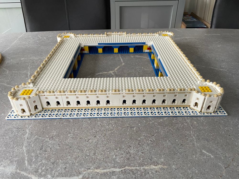 Taj Mahal onderste laag.

LEGO-bouwwerk van een gebouw in de vorm van een vierkante omheining met afgeronde hoeken en een groot open midden. De muren zijn opgebouwd uit witte steentjes met kleine boogvormige ramen. De bovenrand is versierd met ronde beige steentjes en op de hoeken zijn gele details zichtbaar. De structuur staat op een vierkant tegelpatroon in blauw en wit. Binnenin zijn aan de binnenmuur blauw-gele details te zien. Het geheel staat op een grijs tafelblad. 