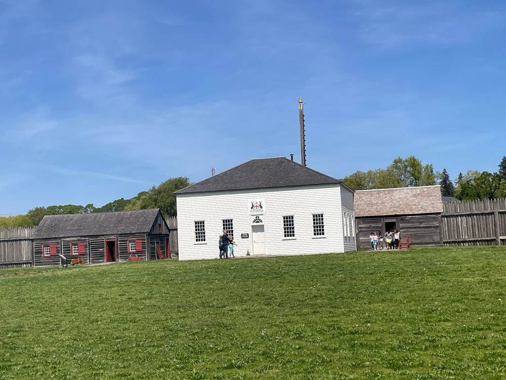 School kids march along a path in front of a historic White House. A fort palisade and a tall pole with a bell is in the background. Other historic buildings are to the left and right.