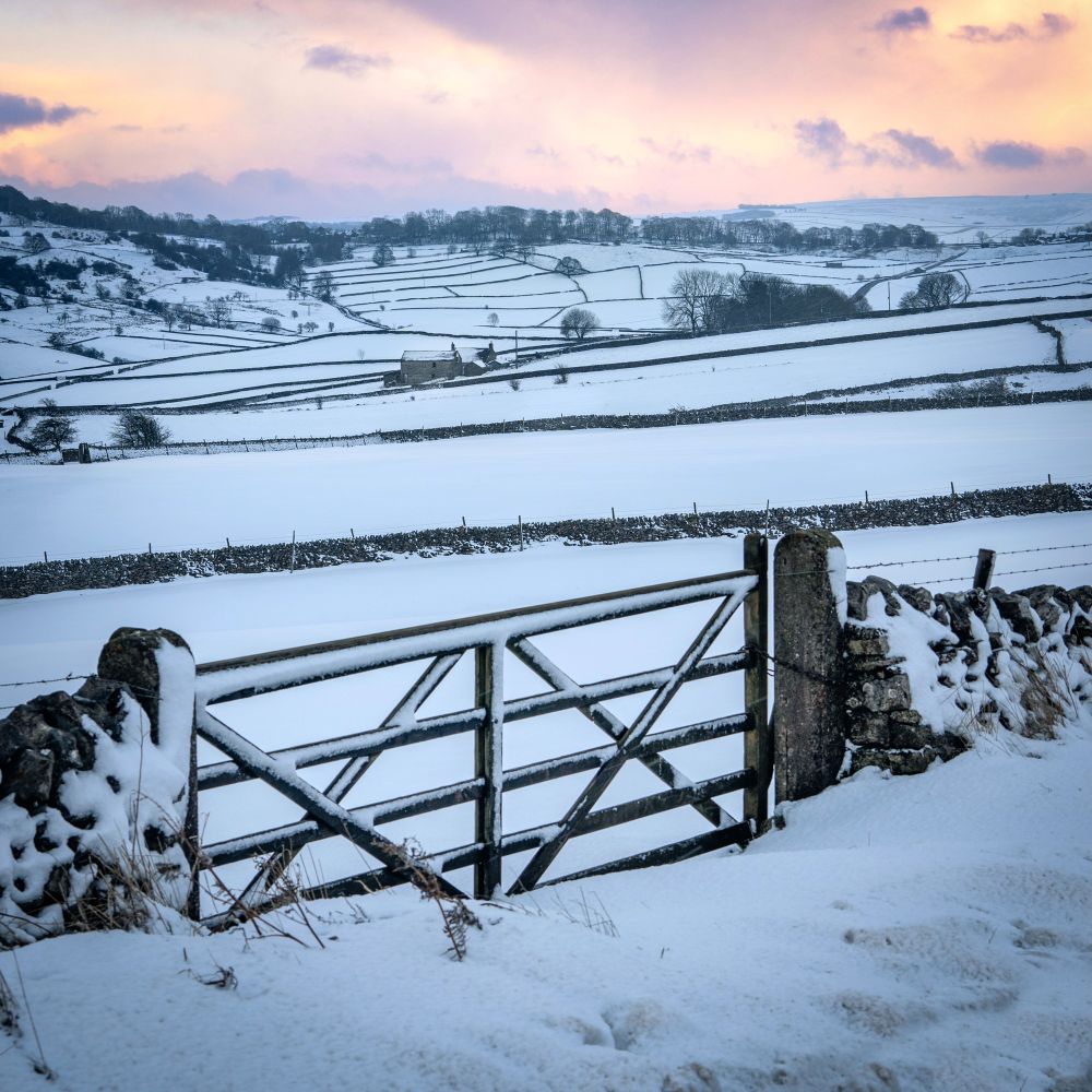 A wooden gate opening to a snow-covered landscape of fields, stone walls and a single farm, leading to distant hills. The sun has just set and the sky is full of pinks, purple and peach hues. 