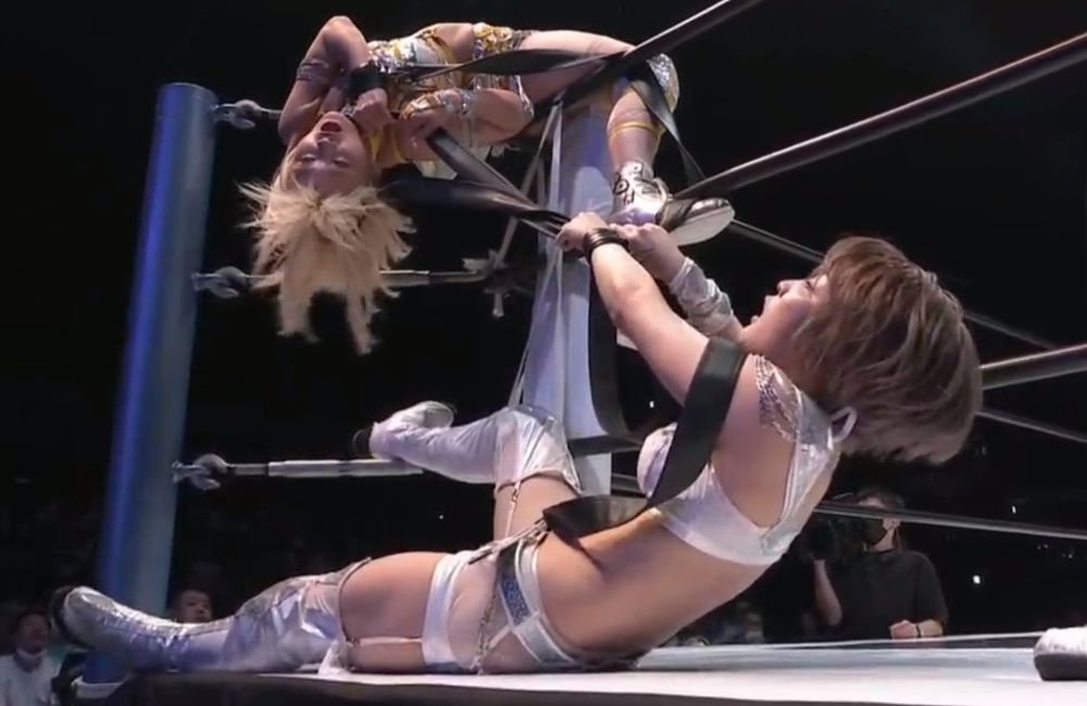 natsupoi is draped over the turnbuckle and saori is lying on the outside apron. saori anou strangles natsupoi by pulling on the strap that connects them together