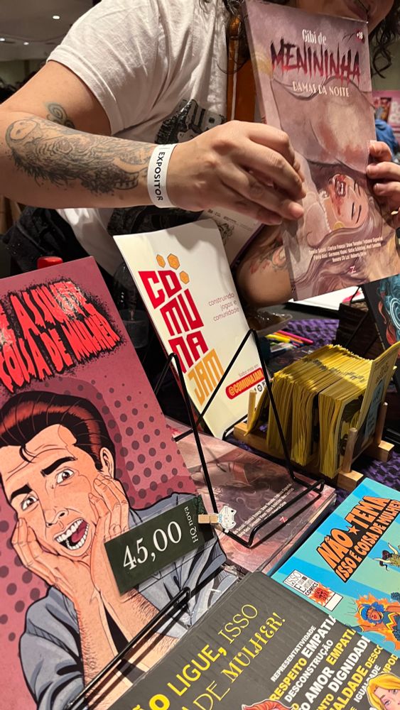 Mesa da Flávia Gasi com vários livros e quadrinhos, e o cartaz da ComunaJam