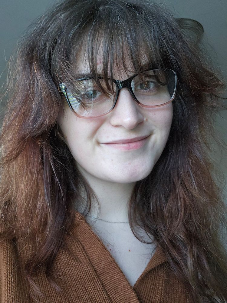 white woman wearing a brown top, necklace, and black glasses with a pink gradient. she has long brown hair. 