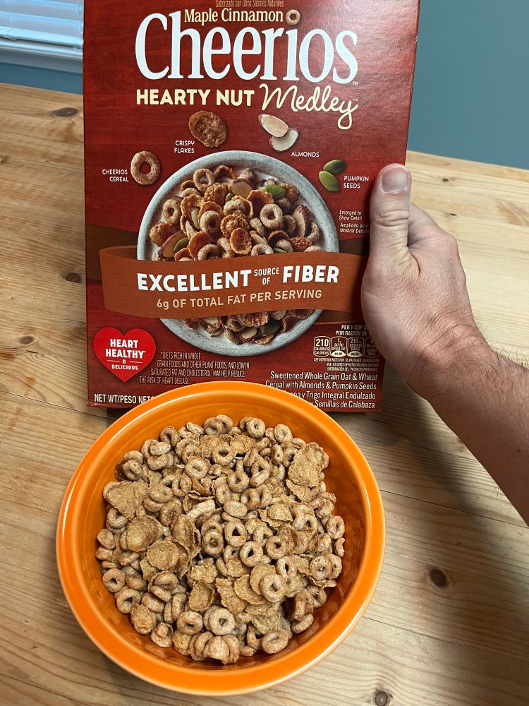 A bowl of hearty nut medley Cheerios cereal without nuts or pumpkin seeds, compared with a box advertising those ingredients