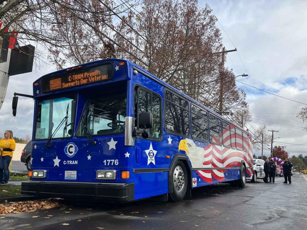 C-TRAN bus 1776 drives along a parade route. The bus is painted in a patriotic color scheme, including a bald eagle and American flag. The headsign on the bus reads C-TRAN proudly supports our veterans.