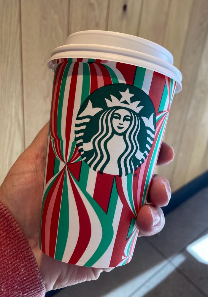 A sultry mermaid with a Satanic star on her crown displays forked Satanic double fins on a cardboard cup adorned with red, white and green flames. The cup contains festive peppermint mocha that is addictive as all Satanic things are. 