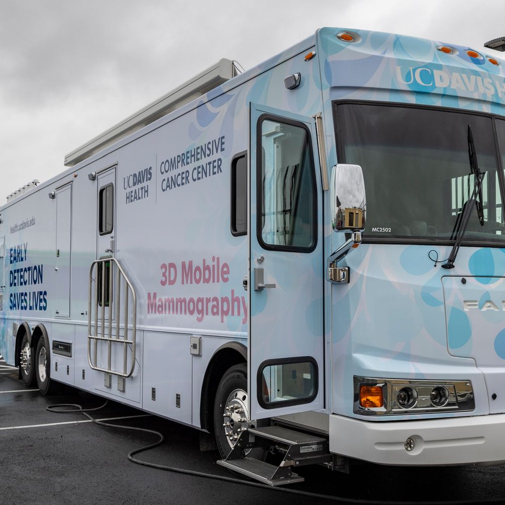 Mobile mammography bus. Large white RV/bus vehicle with signage on the exterior that states "3D Mobile Mammography," "Early Detection Saves Lives," and "UC Davis Comprehensive Cancer Center." The vehicle is parked in a lot with the entry door open for patients.