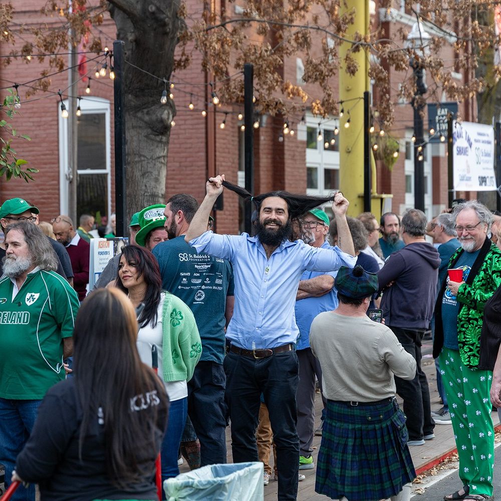 A man pulls his long hair out to the sides to show the length. He is waiting in a line to have his head shaved for St. Baldrick's charity fundraiser.