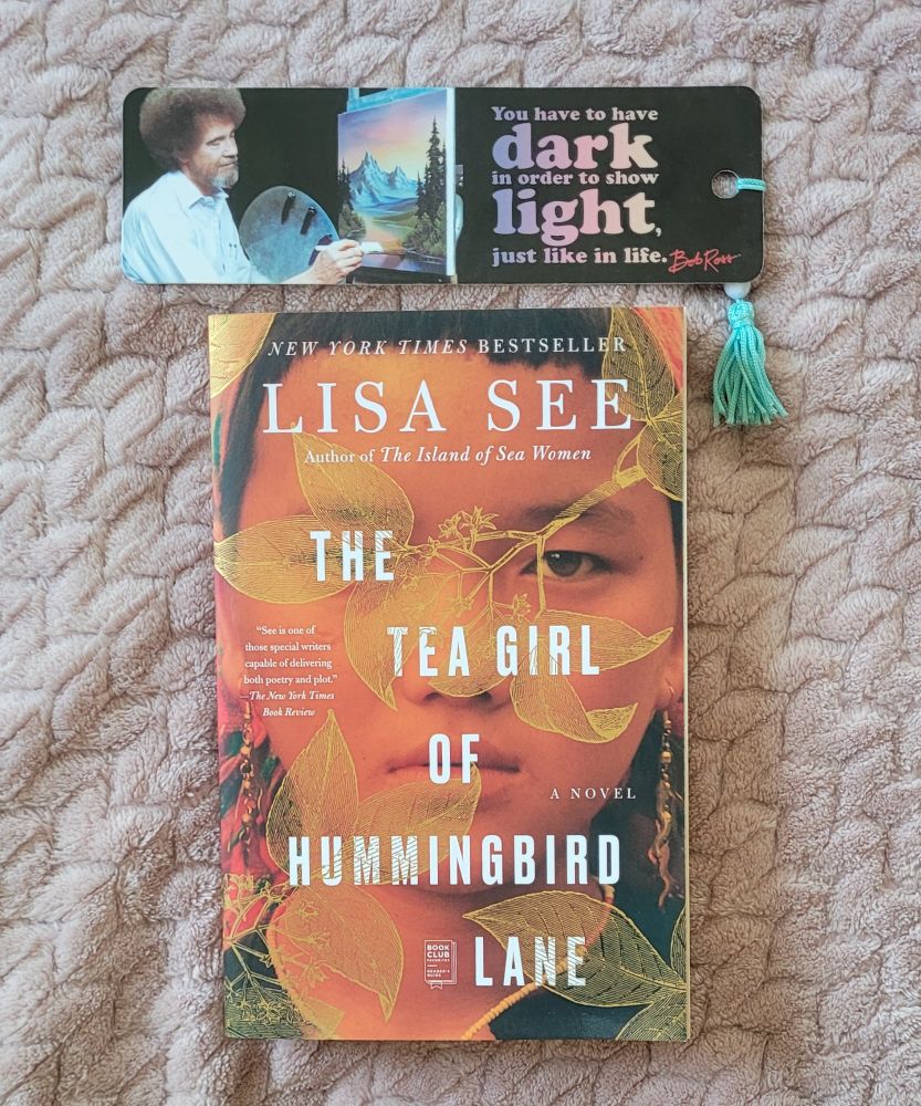 Picture of a bookmark and the book "The Tea Girl of Hummingbird Lane" by Lisa See. The bookmark has a picture of Bob Ross painting and contains the text "You have to have dark in order to show light, just like in life." 