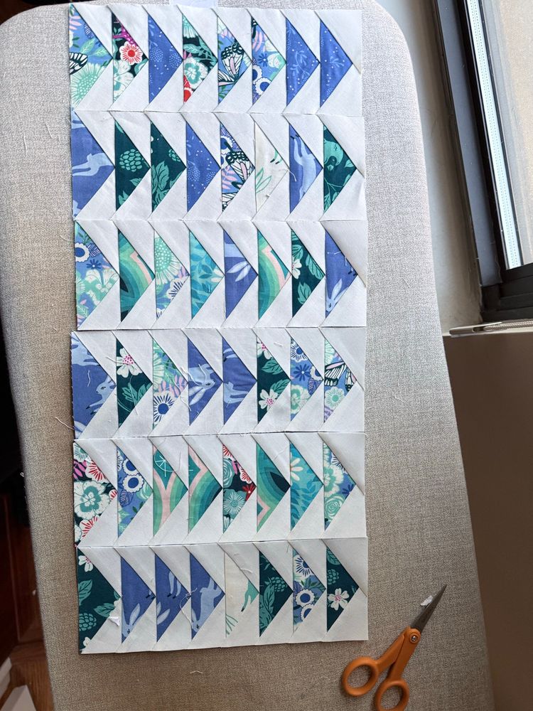 6 pieced fabric strips that will eventually be a quilt. The blocks are comprised of 8 triangles in shades of blue and green surrounded by off white triangles—flying geese blocks. 