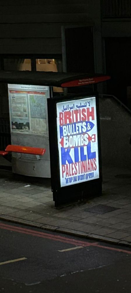 Poster on side of a bus shelter. Text on white background, are red, blue & white highlighted on blue. "British bullets & bombs kill Palestinians"