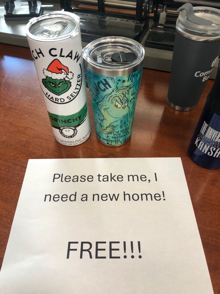 A printer paper sign that says “please take me, I need a new home!! FREE!!!!” in front of two Grinch beverage containers. One is meant to look like a Grinchy white claw