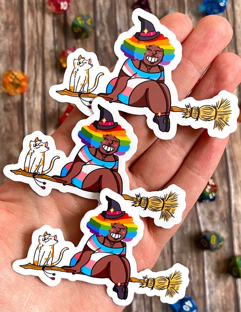 Foto: meine Hand hält mehrere Sticker einer schwarzen Hexe, die auf einem Besen sitzt. ihre Haare sind Regenbogenfarben und ihr Kleid trägt die Trans Pride Farben