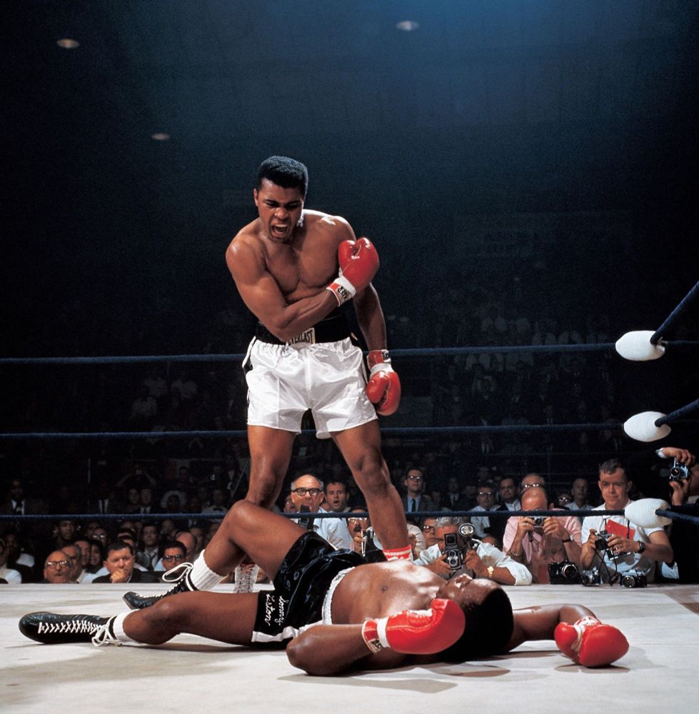 In a boxing ring, Muhammad Ali in white shorts stands over Sonny Liston, who's lying on the ground on his back wearing black shorts. Ali holds up his right hand in a fist. in the background there are photographers and spectators of the match