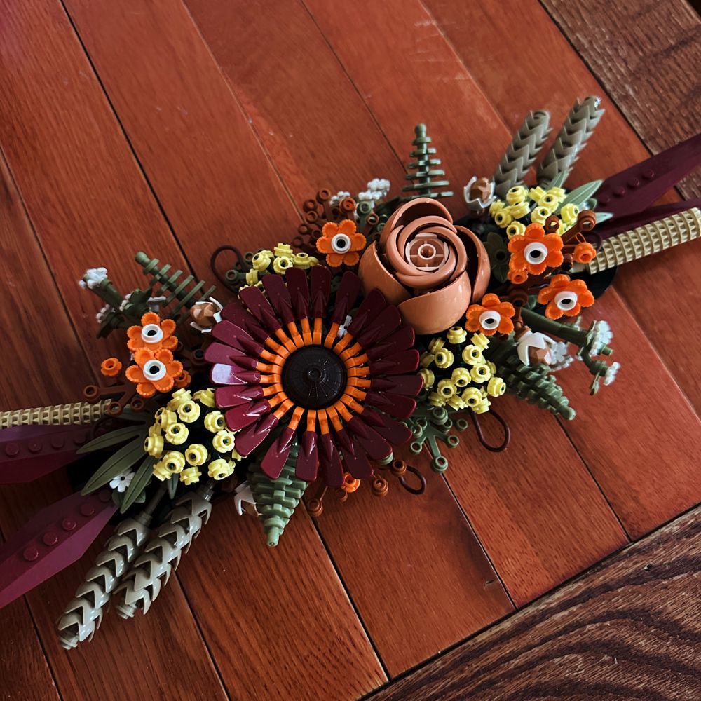 A centerpiece of Lego flowers in autumnal colors, orange, yellow, burgundy, and pale pink, on a wooden table.