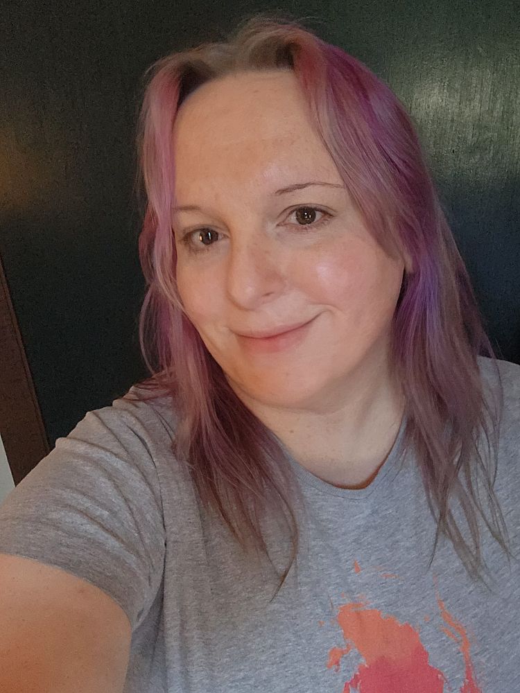 Daria stands in her room in front of a dark green door. She wears a heather gray graphic t-shirt and smiles warmly with her head turned slightly to her right.