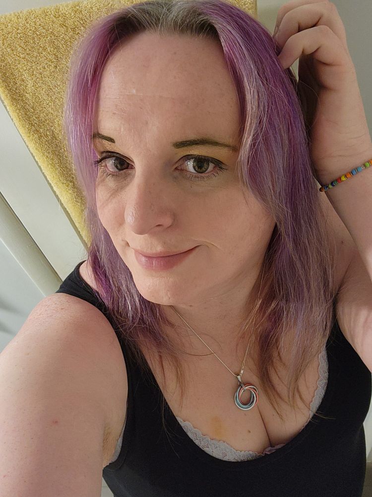 Daria stands in front of a white door with yellow towels hanging on it. She wears a dark blue tank top, a rainbow bead bracelet, and a silver necklace with a pink, blue, and white circle pendant. Her left hand is in her hair at the side of her head and she smiles warmly. 