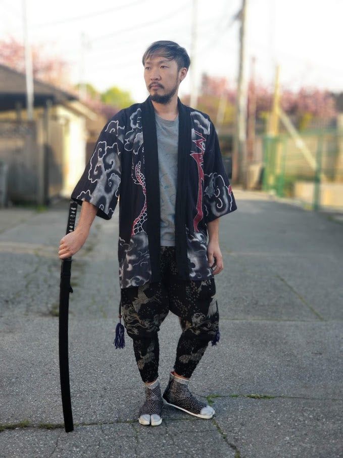 a man in a noragi style jacket, cleft toe boots, grey t-shirt, and patterned pants kind of leaning against a katana 
