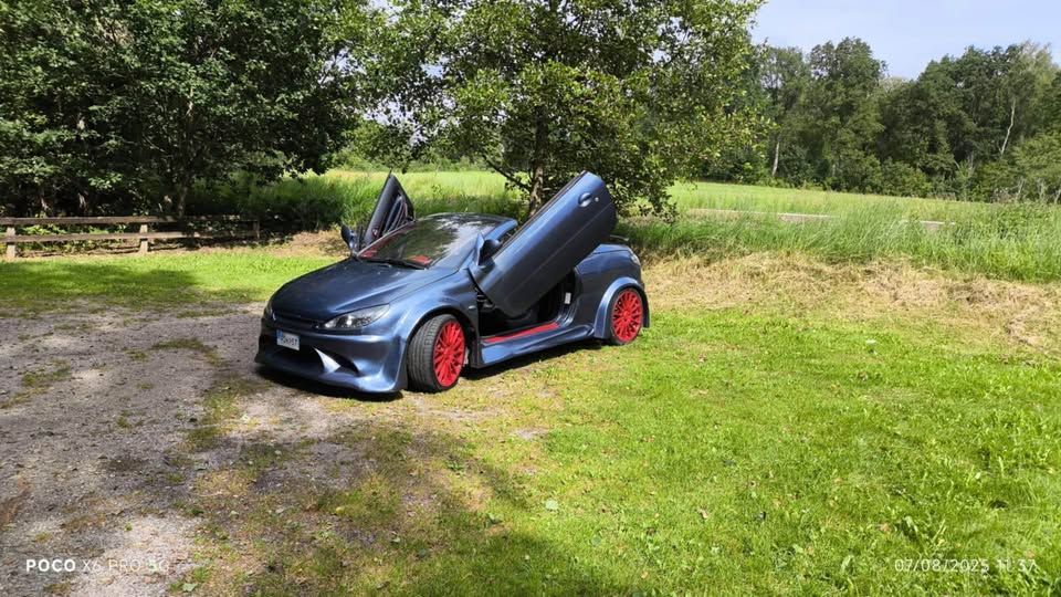 a silver blue peugeot 206cc ricer with its lambo doors open, parked on a field in rural sweden. it has bright red rims and trim.