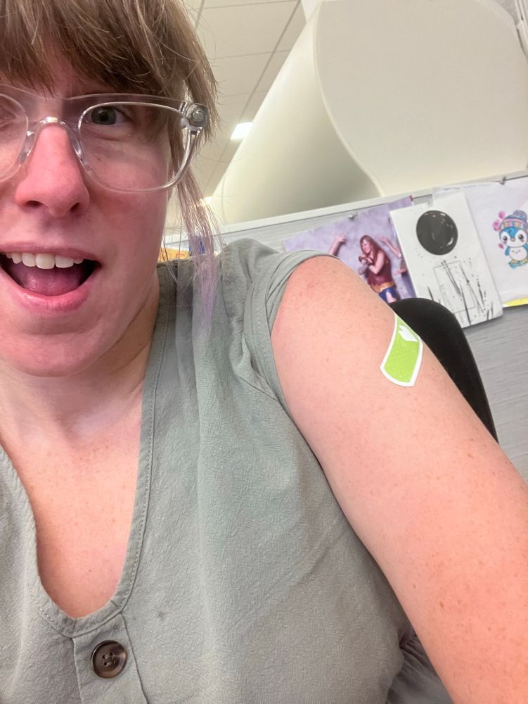 Cat smiles wide with clear framed glasses and a green button up blouse with a green cvs heart bandaid on her bare shoulder