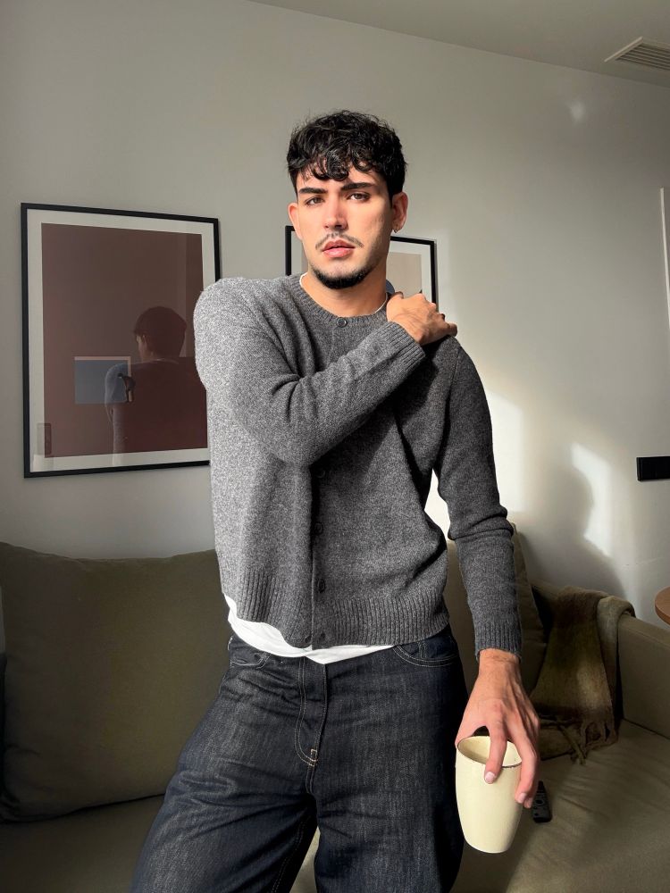 A picture of a handsome man indoors wearing a cardigan and holding a cup of coffee. Very aesthetic.