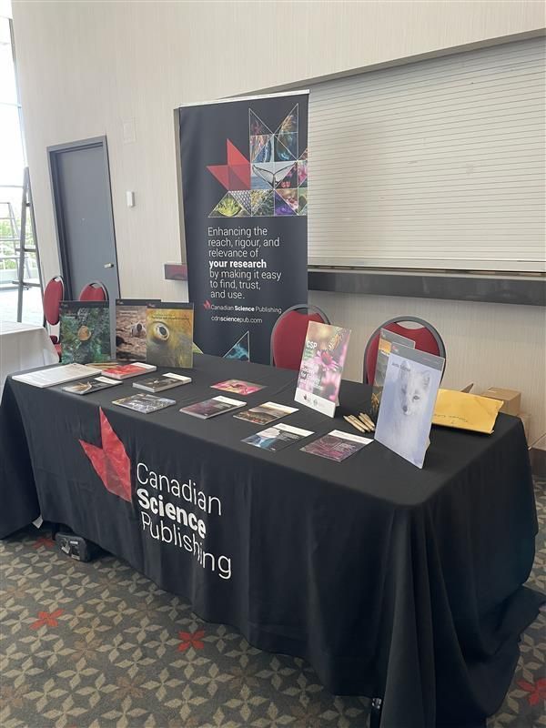 A conference booth with a black Canadian Science Publishing tablecloth. Various journal summary cards and covers are on display. A banner behind the table reads "Enhancing the reach, rigour, and relevance of your research by making it easy to find, trust, and use."