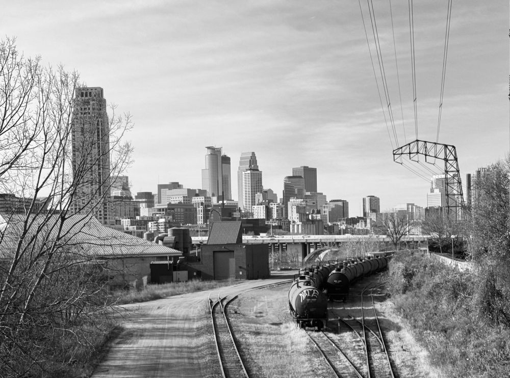 The black and white image shows a cityscape with a variety of skyscrapers in the background and train tracks in the foreground. The skyline features modern high-rise buildings with reflective glass surfaces and older structures with more traditional architecture. In the foreground, there are parallel train tracks leading into a series of black tanker rail cars, which are lined up. On the left, there are industrial buildings. To the right, a patch foliage and bare trees runs alongside the tracks. Overhead, power lines are supported by a tall transmission tower on the right side of the image. 