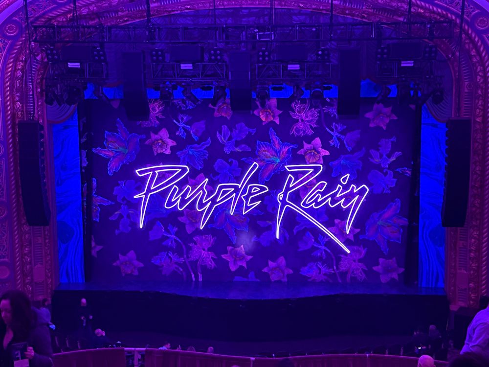The stage right before Purple Rain - The Musical. It features a backdrop of flowers, the words “purple rain” and everything is lit up in purple. 
