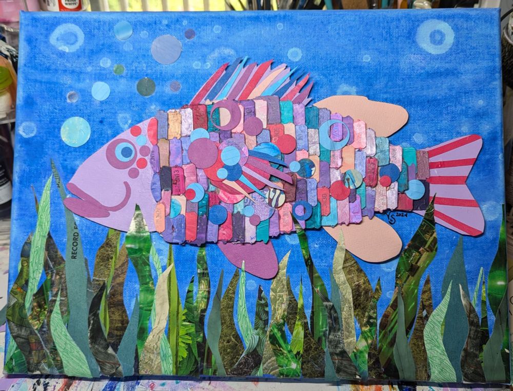 A mixed media piece with a large colourful fish swimming amongst some reeds. Made mostly out of recycled paper, glued to canvas.