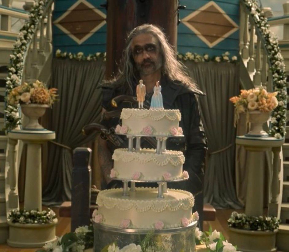 Edward Teach in season 2 episode 1 of Our Flag Means Death, wearing his Blackbeard leathers with black kohl across his eyes and mouth, standing in front of a large wedding cake on the deck of a ship during a raid with a lifeless expression