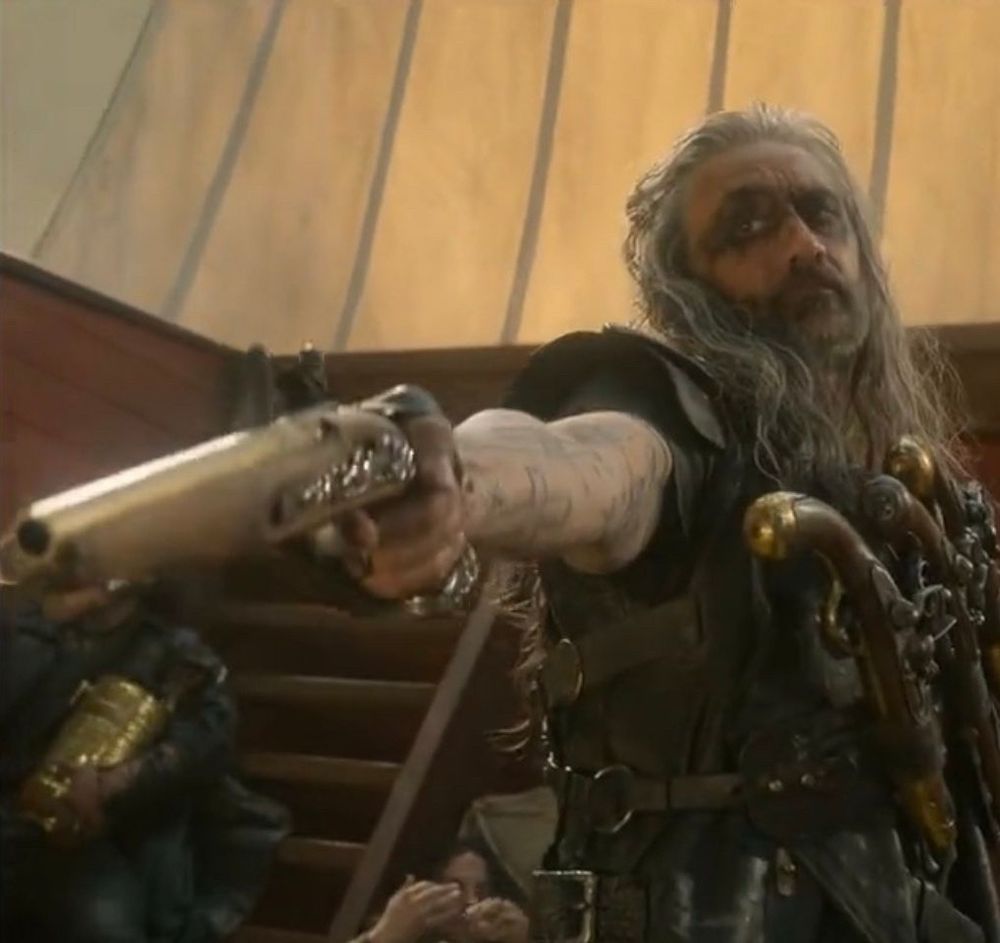 Edward Teach in season 2 episode 1 of Our Flag Means Death, wearing his Blackbeard leathers with a bunch of pistols strapped to his chest and black kohl across his eyes and mouth, pointing his pistol at a wedding guest out of frame during a raid with a lifeless expression