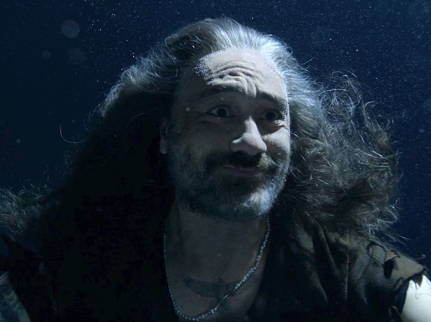 Edward Teach in season 2 episode 3 of Our Flag Means Death, wearing a black t-shirt and pearls, looking at Stede out of frame underwater in the Gravy Basket with a loving smile